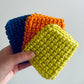 Three crochet squares in blue, orange, and green held by a hand against a light gray background.