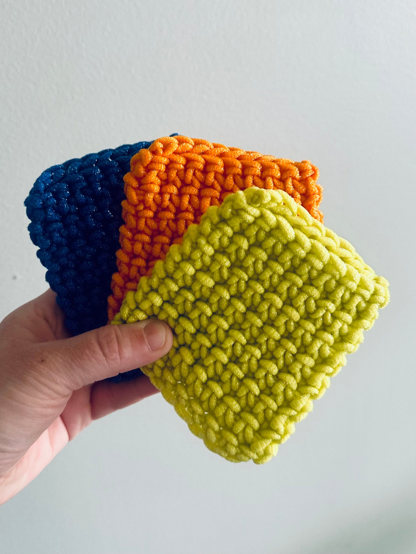 Three crochet squares in blue, orange, and green held by a hand against a light gray background.