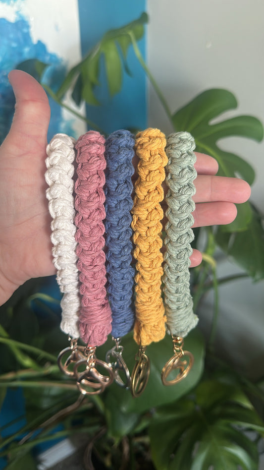 Hand holding five colorful crocheted keychains with matching keychains below, against a blurred green plant background.