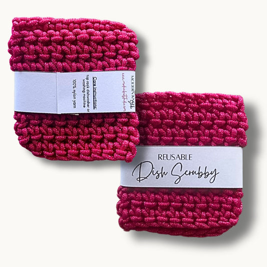 Two hot pink crochet dish scrubbies with labels, one labeled 'REUSABLE DISH SCRUBBY' and the other with a care instruction label.